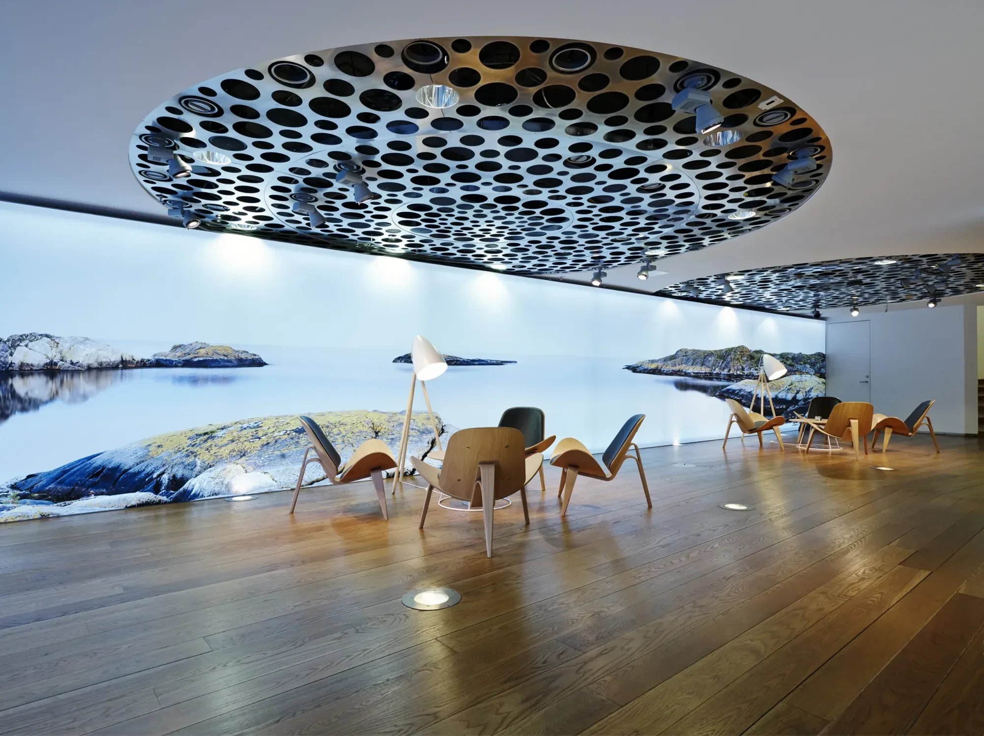 Modernt loungeutrymme med trägolv, designmöbler och stora perforerade takinstallationer. Längs väggen löper en panoramabild av ett lugnt kustlandskap med klippor och hav.